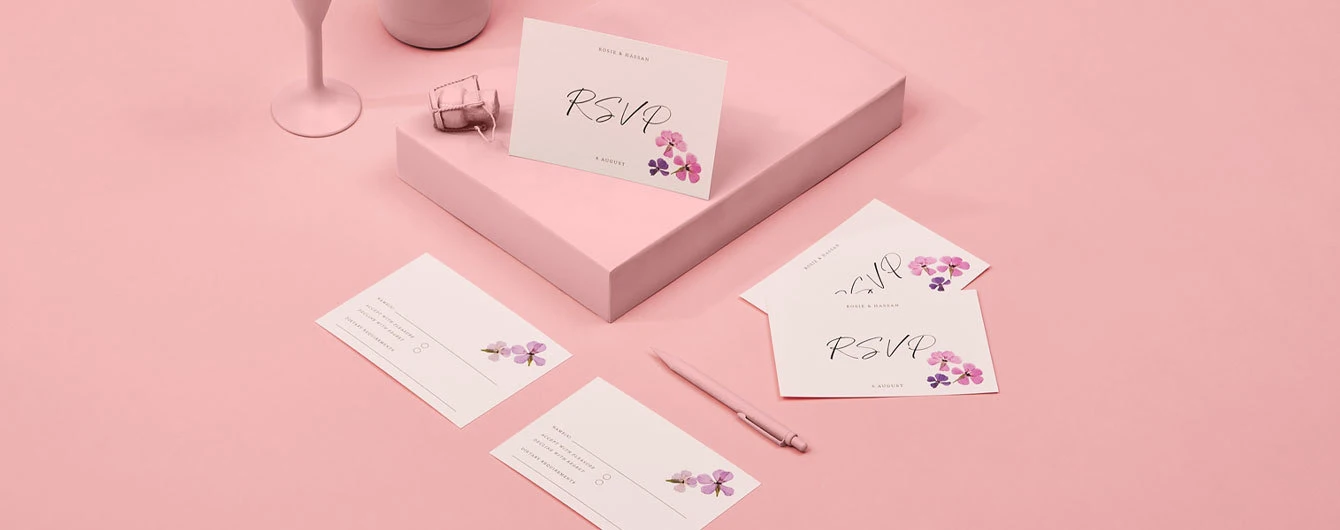 A collection of elegant pink wedding RSVP cards decorated with small purple and pink flowers, artfully displayed with a matching pen, wine glass, and champagne cork on a dusty pink background.