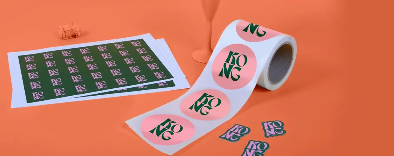 A roll of large, round pink stickers printed with the green text ‘KONG’ alongside flat sheets of smaller rectangular stickers and individual die-cut labels, all arranged on a solid orange background.