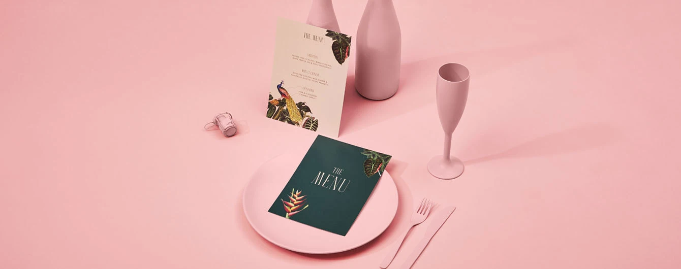 A pink table setting displaying a dark green wedding menu card on a dinner plate and a matching cream menu card upright, both featuring elegant serif text and tropical illustrations.