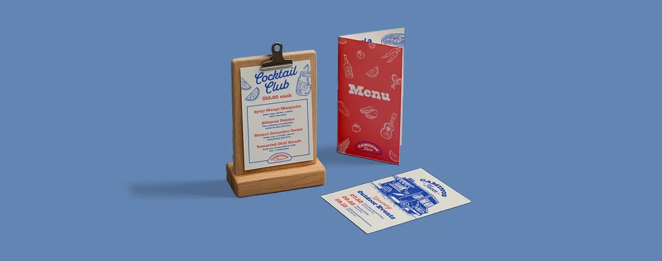 A restaurant table display on a blue background, featuring a 'Cocktail Club' menu on a wooden clipboard stand, a folded red food menu, and a flyer listing upcoming outdoor events.