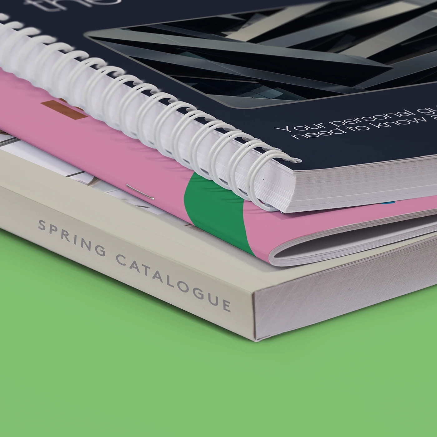 A close up showing a stack of three different types of Catalogues — a perfect bound "Spring Catalogue" on the bottom, a pink saddle stitch Catalogue in the middle, and a navy spiral bound Catalogue on top.