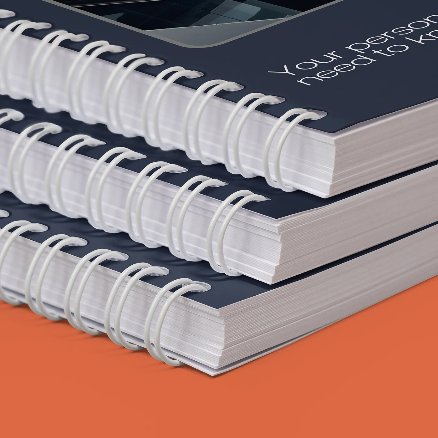 A close-up shot shows a stack of three navy blue Catalogs with prominent white double-loop wire bindings resting on a solid orange background.
