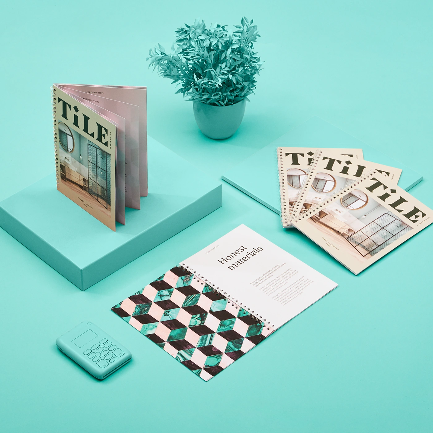 Three Spiral Bound Catalogues shown both open and fanned out, arranged on a turquoise surface with a matching plant pot and electronic device.