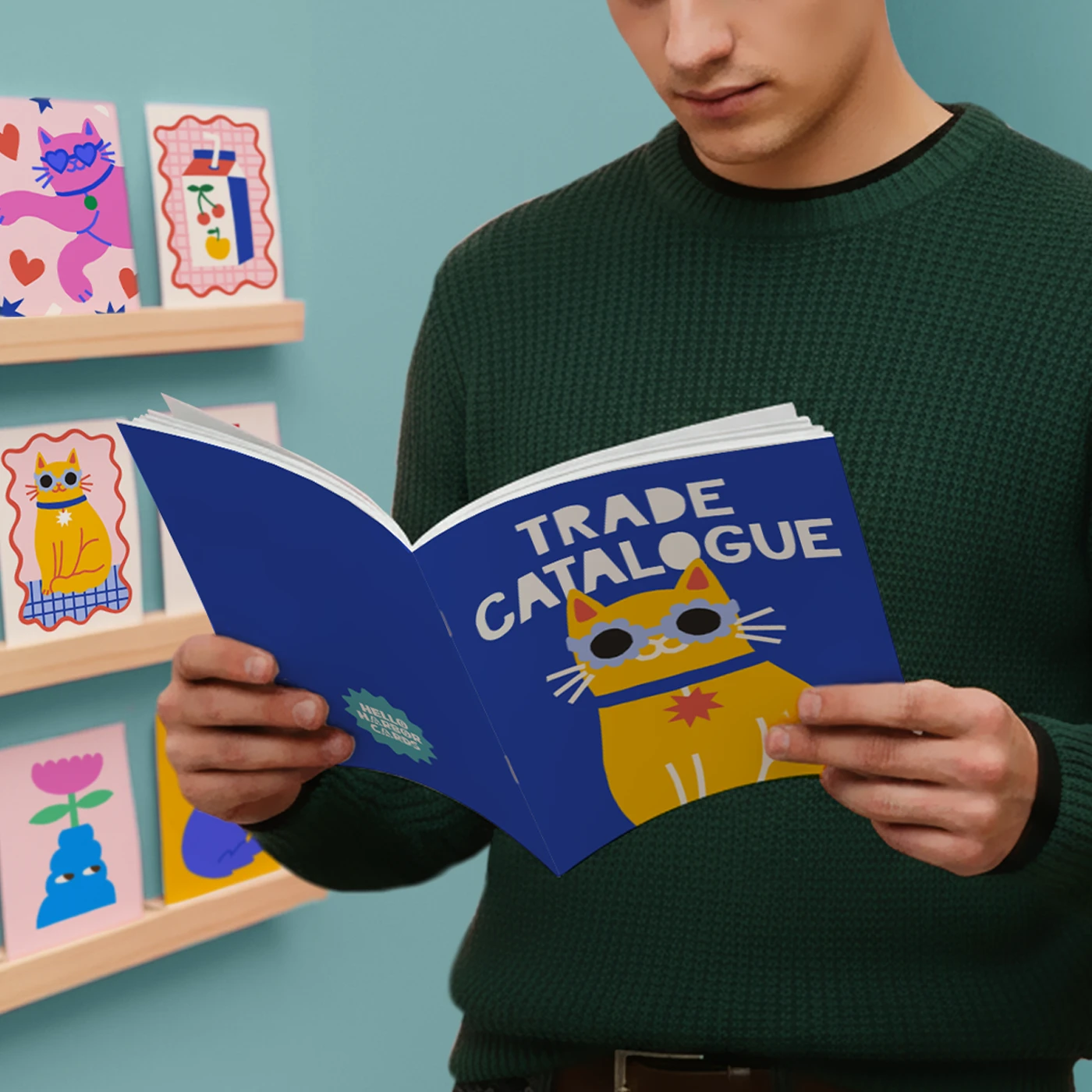A person holds open a blue saddle stitch trade Catalogue featuring a cartoon yellow cat wearing sunglasses, with illustrated cards displayed on shelves in the background.