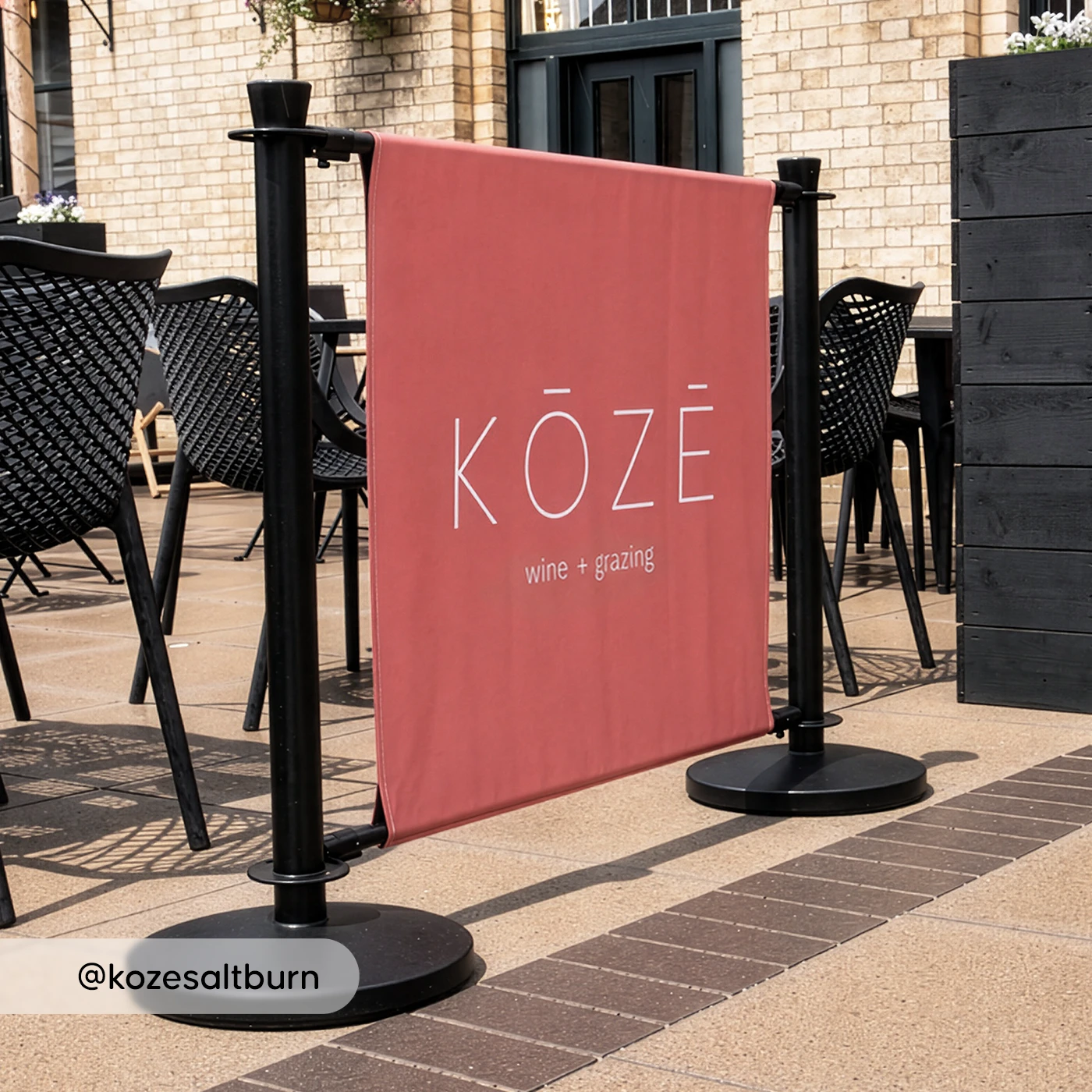 Red KOZE cafe barrier displayed outside a seating area with tables and woven chairs