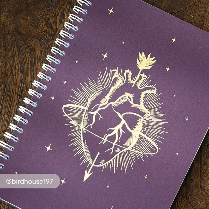 An gold foil anatomical heart pierced by an arrow and surrounded by radiant lines and small stars on a purple Spiral Bound Journal cover.