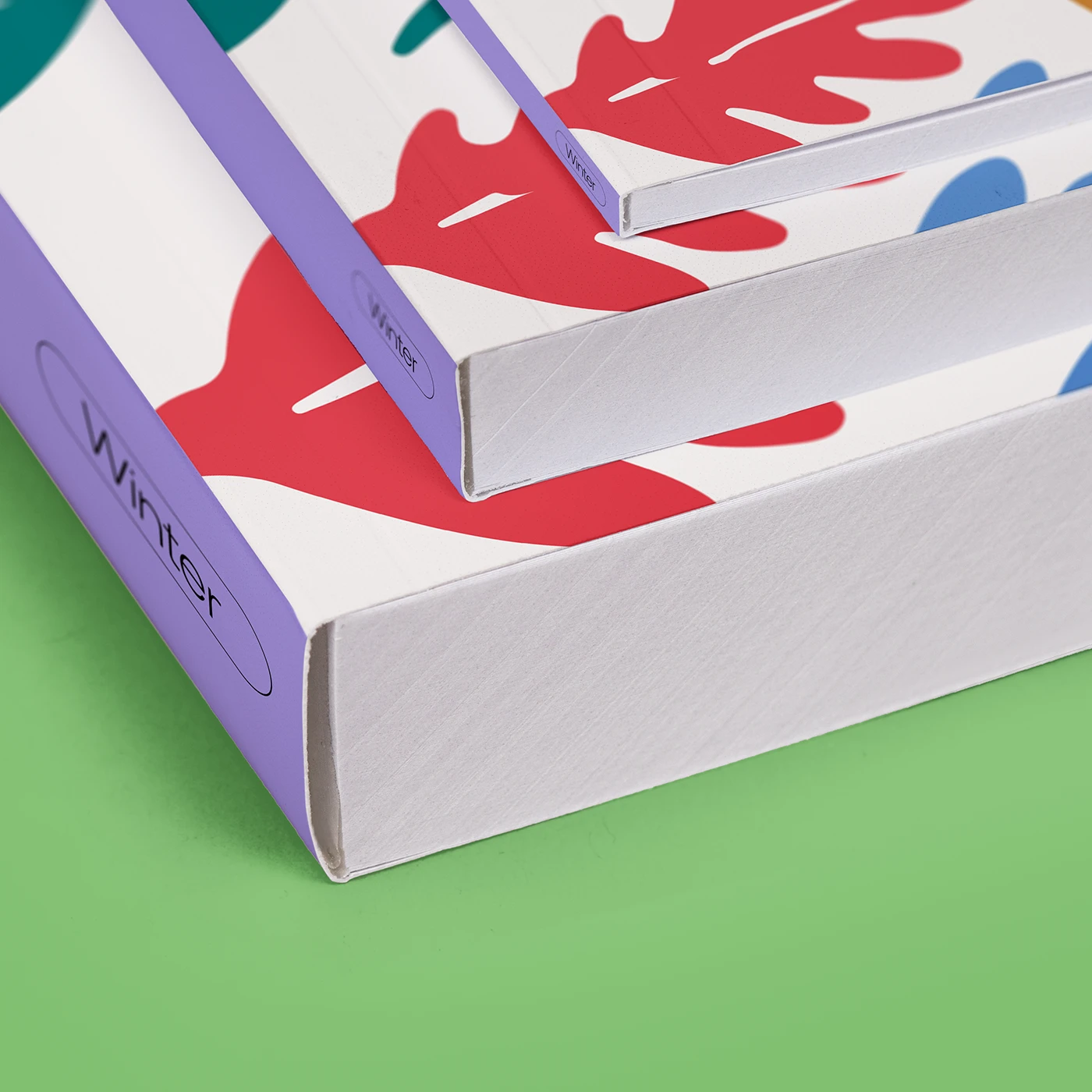 A stack of three Perfect Bound Books of varying thicknesses and page counts featuring purple spines labelled "Winter" and covers decorated with red and blue graphic shapes on a light background.