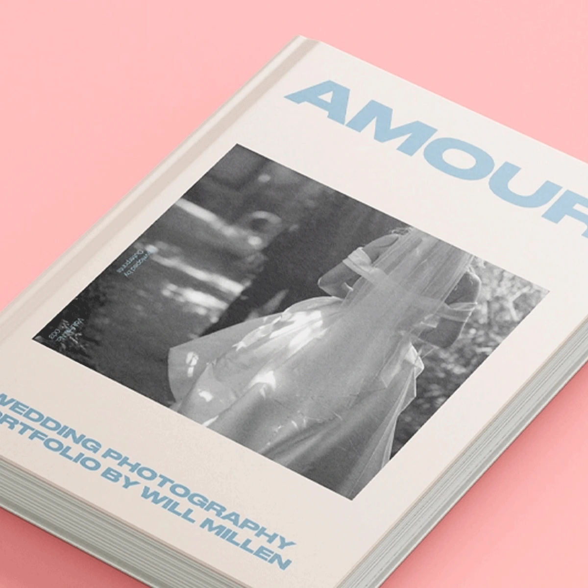 Angled view of a white Hardback Book titled "AMOUR" featuring a black-and-white photograph of a bride and light blue text reading "Wedding Photography Portfolio by Will Millen,"
