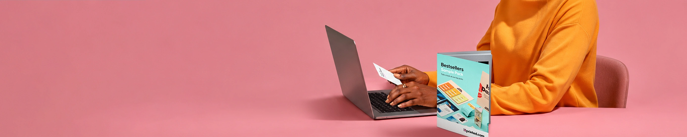 A person in a bright orange jumper works on a laptop at a desk against a pink background, holding a small white card next to a standing printed booklet titled "Bestsellers Sample Pack".