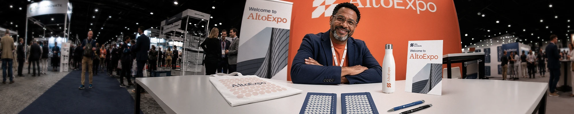 A smiling man in a navy blazer sits at an AltoExpo exhibition booth featuring branded stationery, a tote bag, a water bottle, and promotional banners.