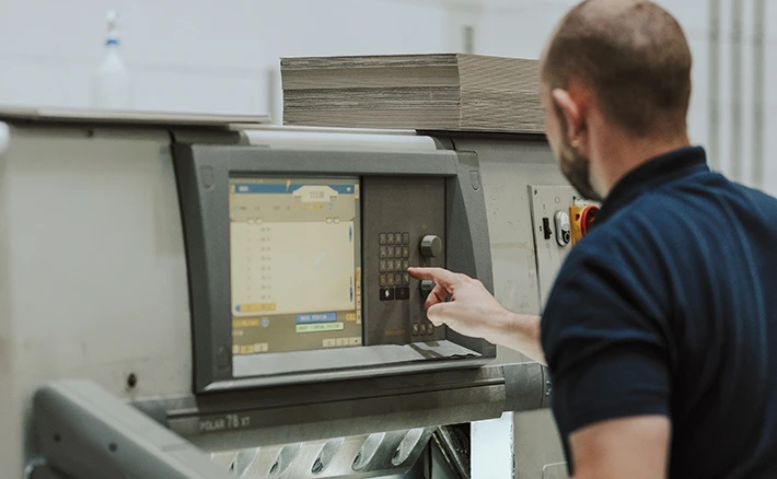 A print technician operates the digital control panel of a large paper-cutting machine stacked with card.