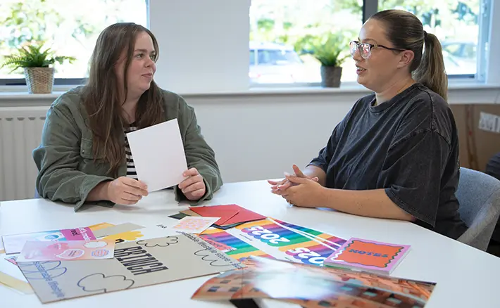 Two women sit opposite each other at a bright office table covered with colorful stationery design samples, discussing ideas as one holds a blank card.