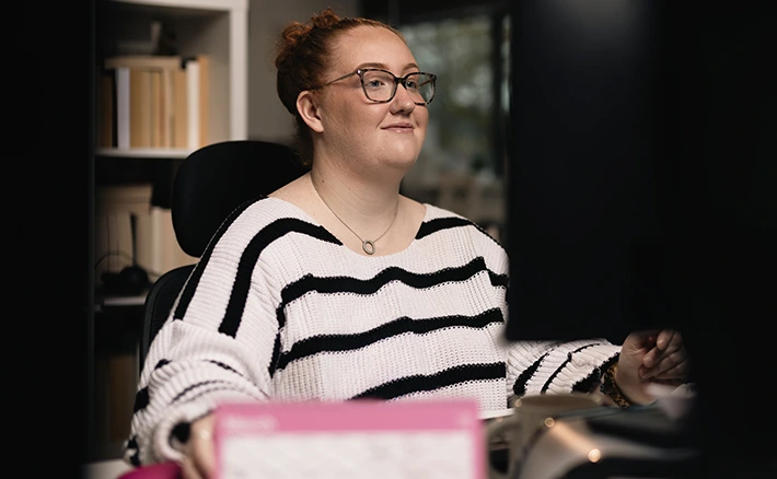 A woman with glasses and red hair tied up in a bun smiles while looking at her computer monitor in an office setting.