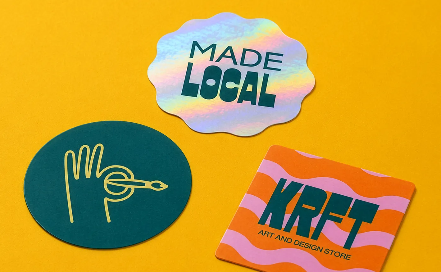 Three individual custom stickers laid out on a yellow background: a scalloped holographic sticker reading "MADE LOCAL", a circular dark green sticker with a hand illustration, and a rectangular orange and pink sticker for "KRFT Art and Design Store".