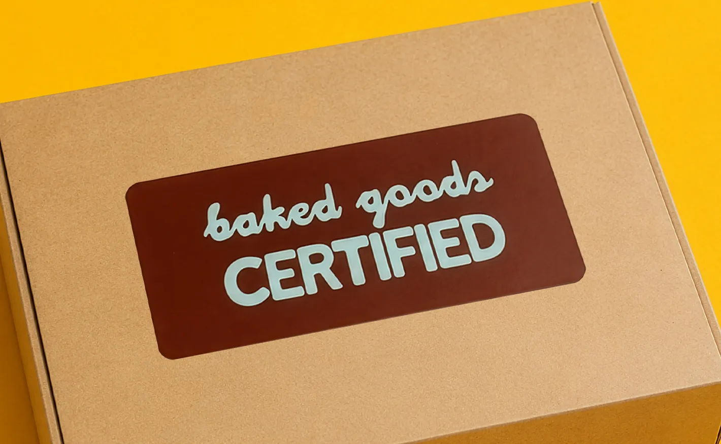 A large brown rectangular sticker with light blue text reading "baked goods CERTIFIED" fixed to the centre of a cardboard packaging box.