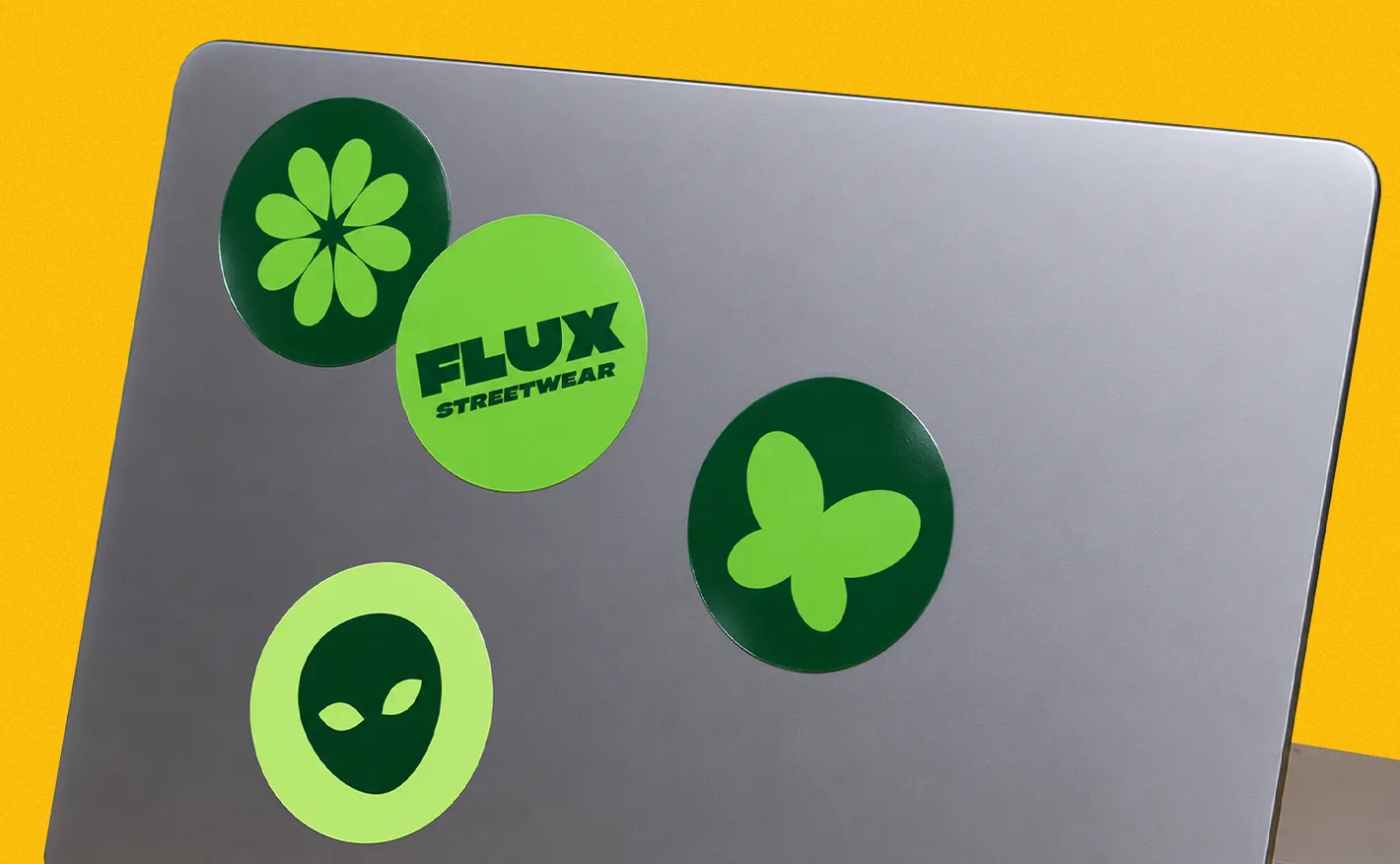 Four circular green vinyl stickers, featuring graphic designs like a flower, an alien head, a butterfly, and the text "FLUX STREETWEAR", decorated across the corner of a grey laptop lid.
