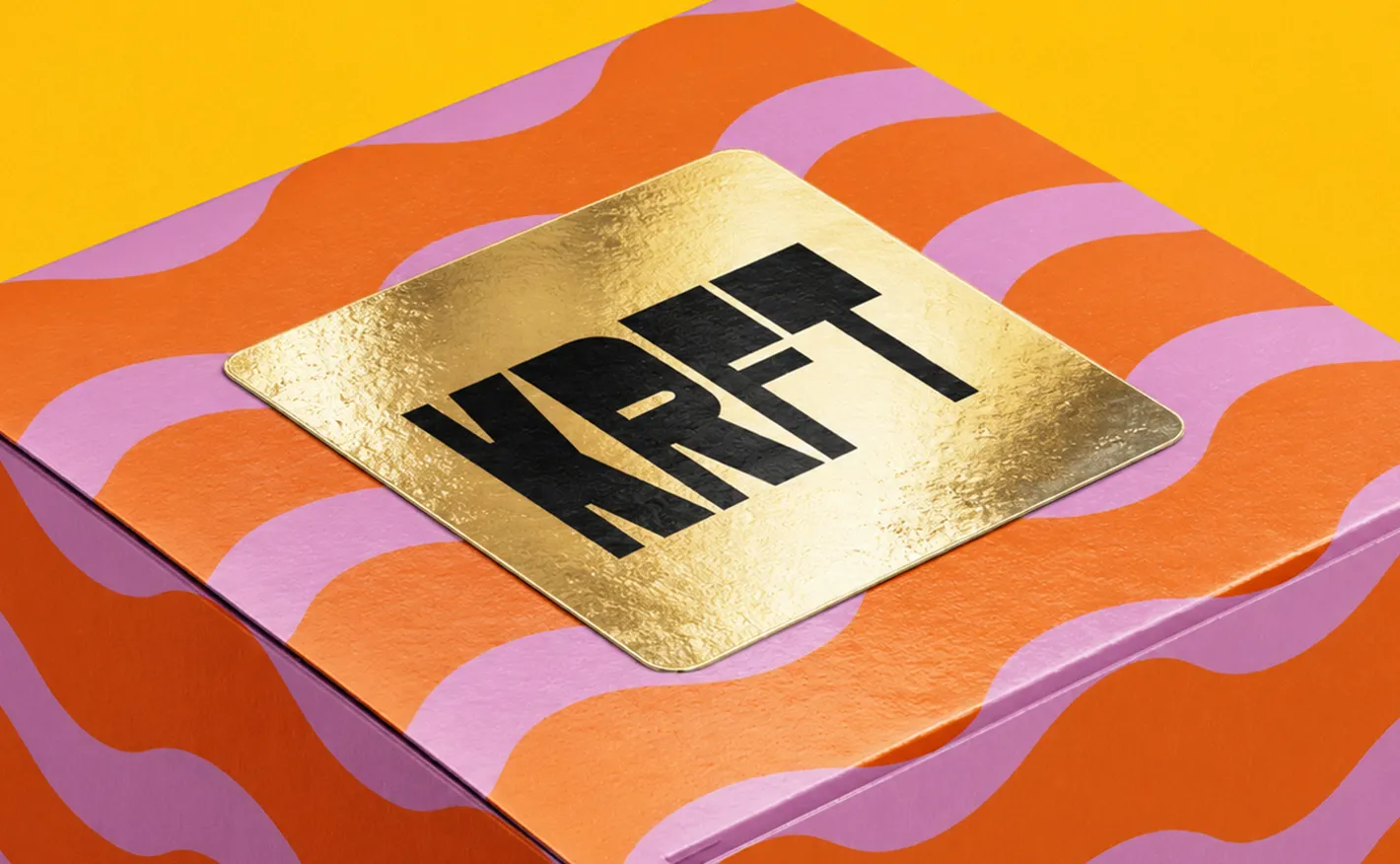 A square metallic gold sticker printed with black "KRFT" branding, sealing the top of a pink and orange wavy-patterned cardboard box.