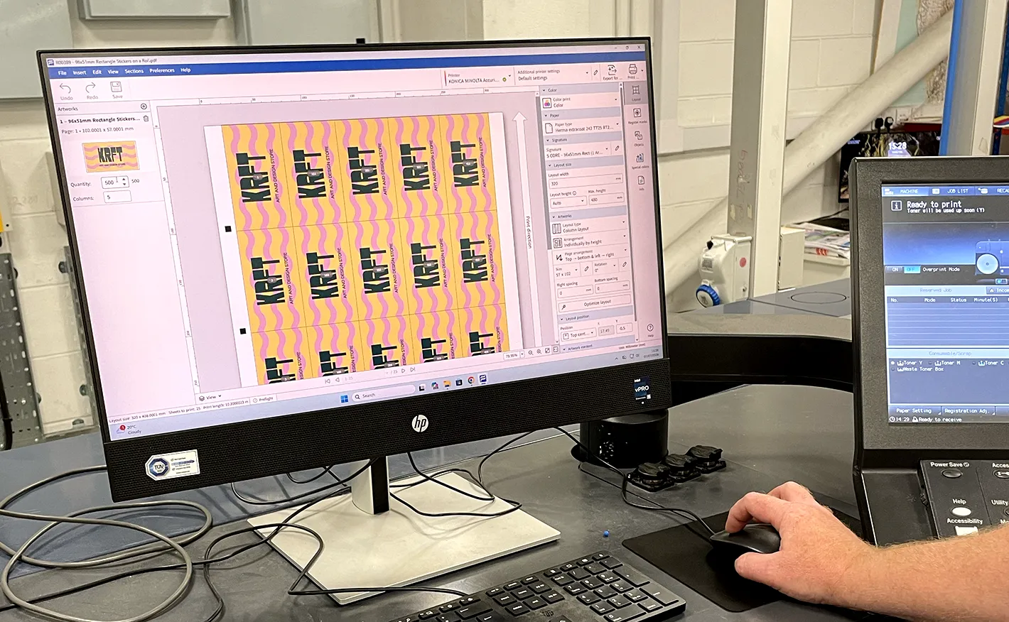 A prepress operator using a computer mouse to adjust the print layout of a batch of yellow and pink "KRFT" stickers on a desktop monitor.