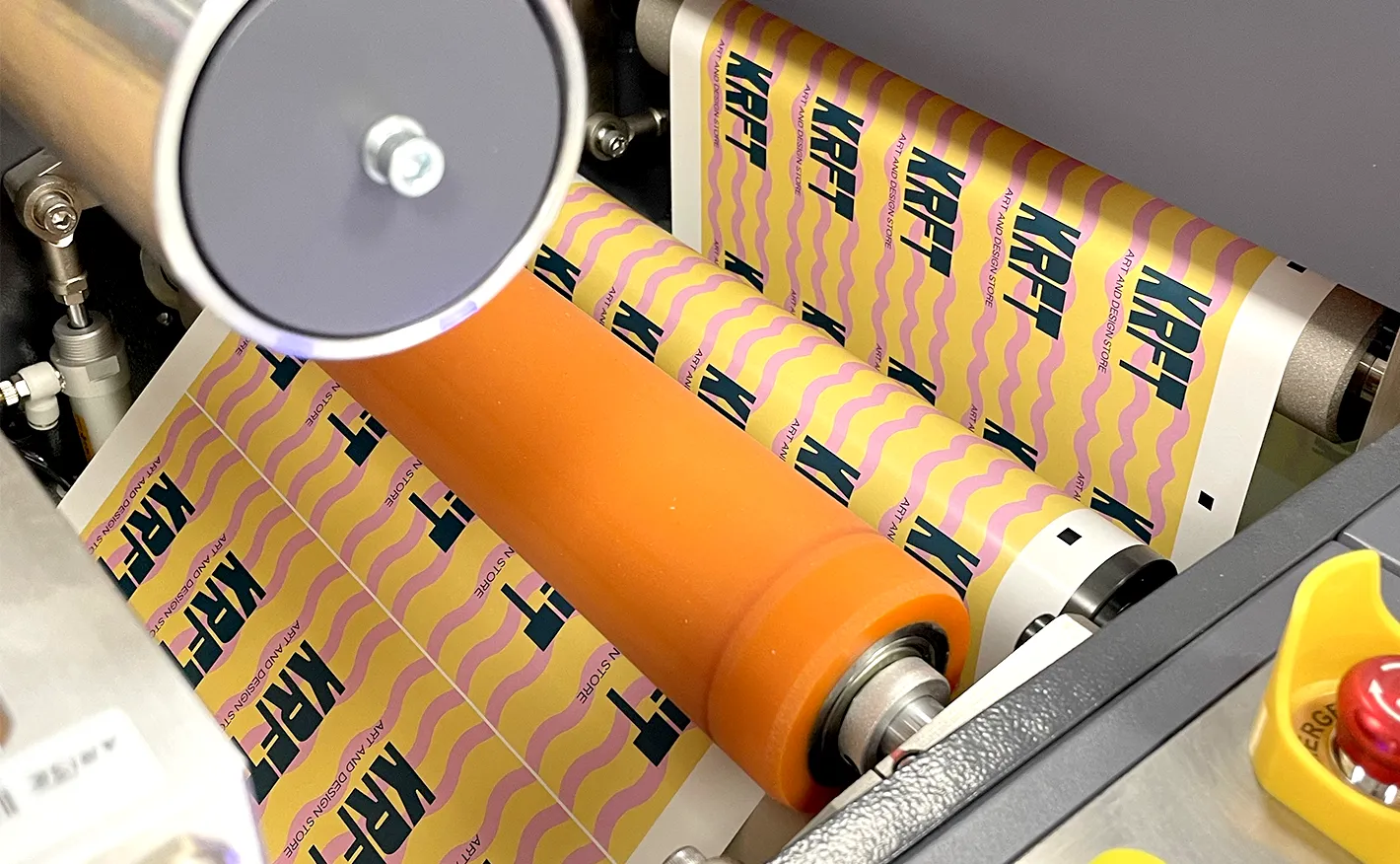 A close-up of a printing press feeding a continuous roll of pink and yellow "KRFT" stickers under an orange pressure roller.