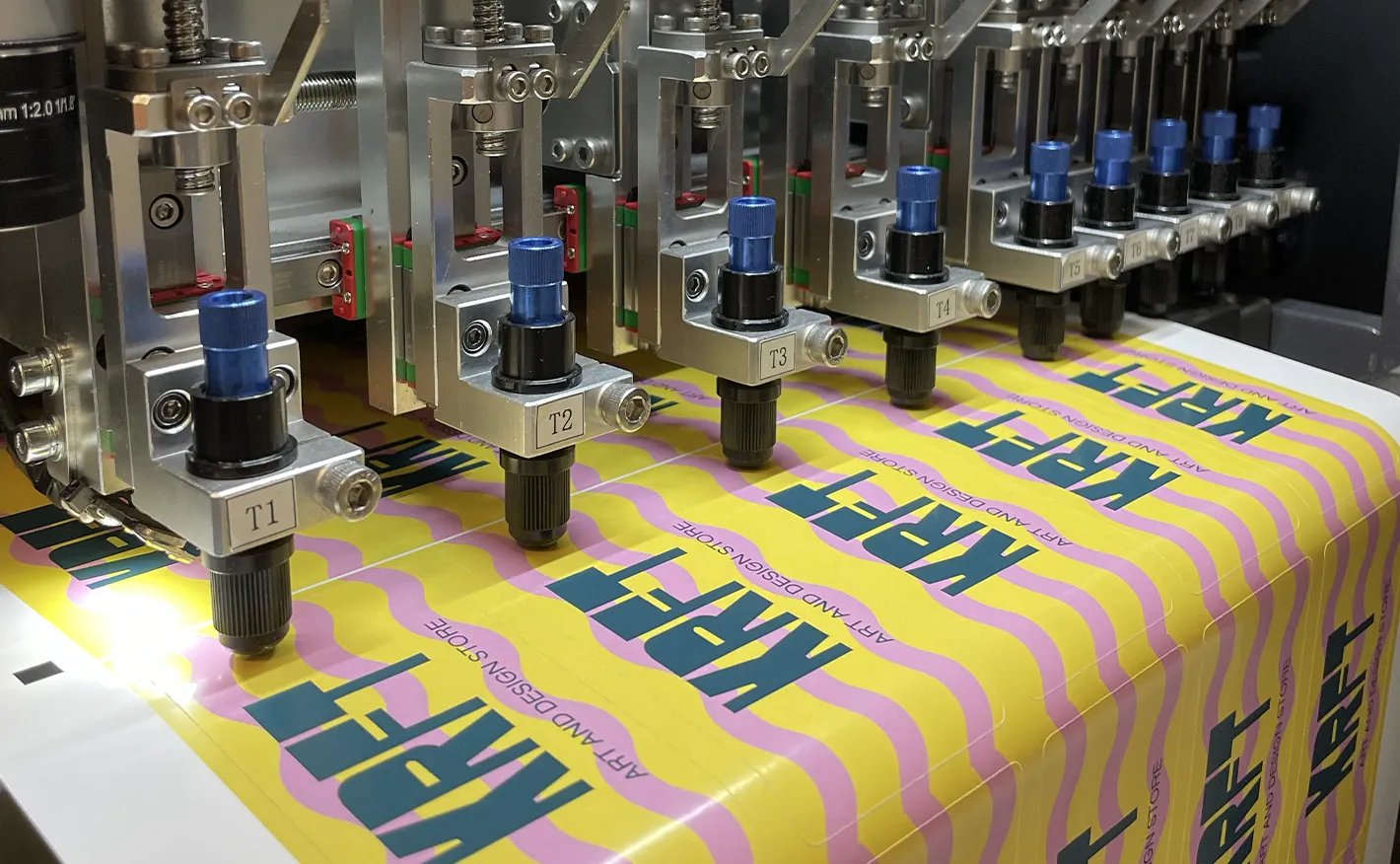 An automated cutting machine precision-cutting a sheet of yellow and pink wavy stickers featuring the "KRFT Art and Design Store" logo.
