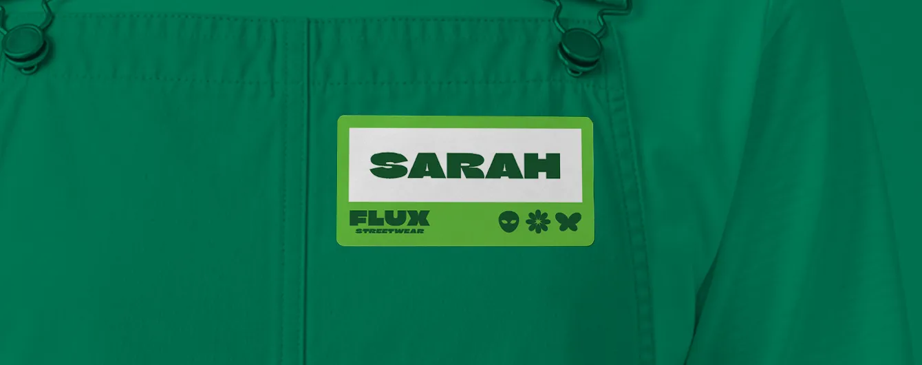 A green and white name badge sticker printed with "SARAH" and "FLUX STREETWEAR" attached to the bib of a green apron.