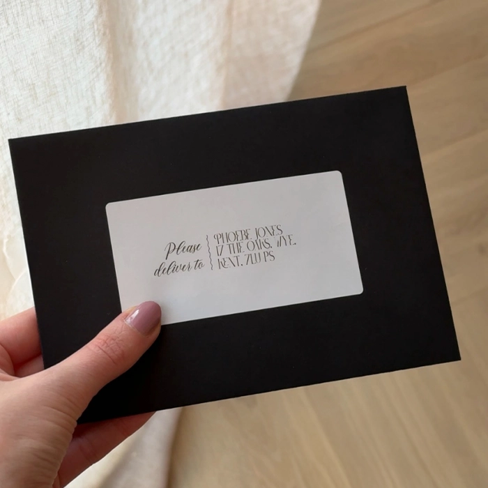 A hand holds a black Envelope with a rectangular white Address Label reading, "Please deliver to: Phoebe Jones, 17 The Oaks Way, WA12 34BS."