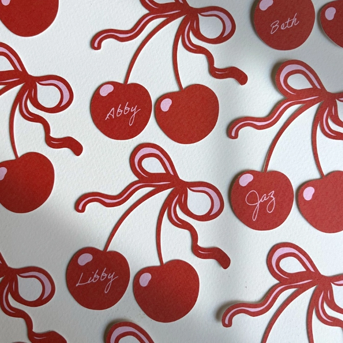 Bespoke cherry-shaped Place Cards tied with pink-outlined red bows, handwritten with names like "Abby," "Libby," "Jaz," and "Beth" in white cursive.