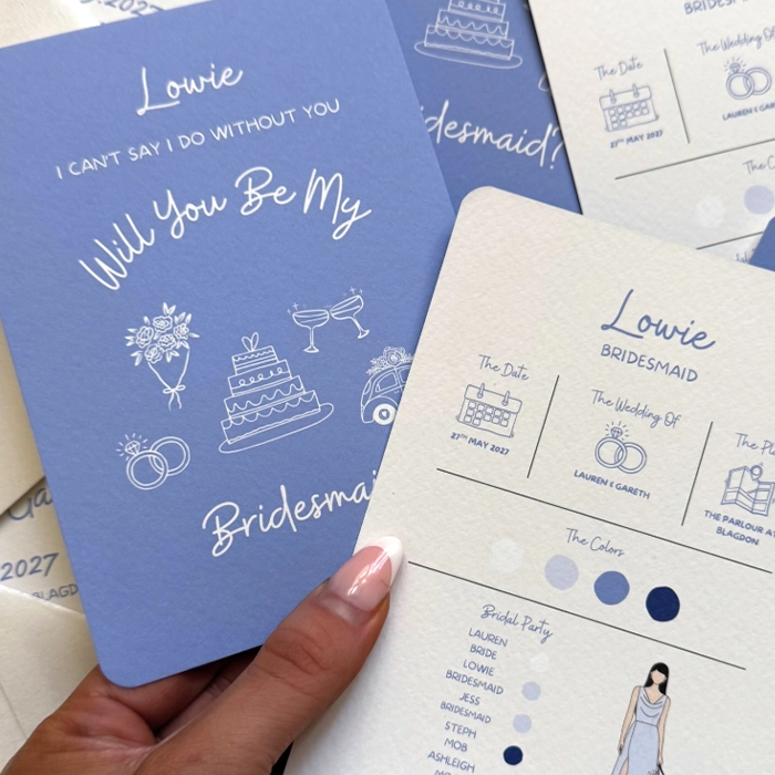 A hand holding a periwinkle blue "Will You Be My Bridesmaid" card alongside a matching white information card displaying wedding details and colour swatches.