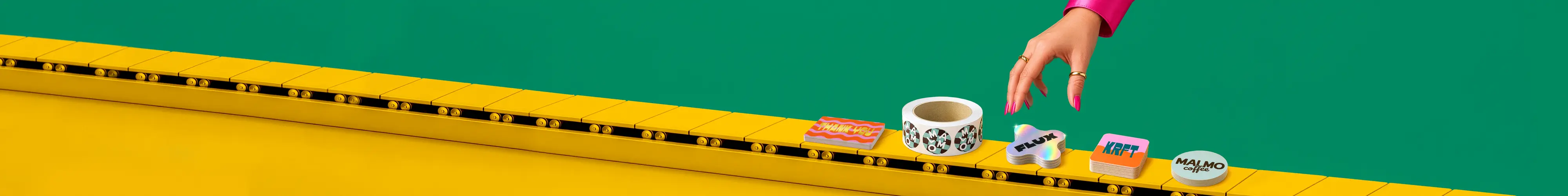 A hand with pink nails reaches towards a selection of custom stickers and a sticker roll displayed along a yellow conveyor track against a green background.