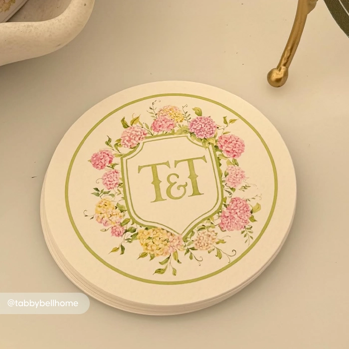 A stack of round Paper Coasters featuring a floral wreath of pink and yellow hydrangeas surrounding a monogram that reads "T&T".