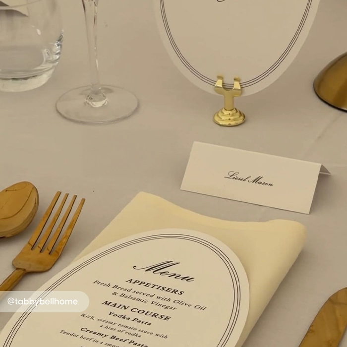 An elegant table setup featuring an arched menu card resting on a cream napkin, a folded Place Card for "Liesel Mason," and a round card in a gold stand.