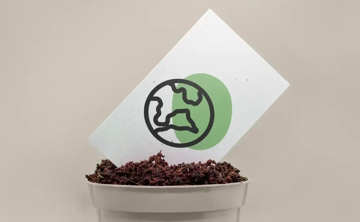 A white card printed with a green globe icon resting inside a small plant pot filled with soil against a grey background.