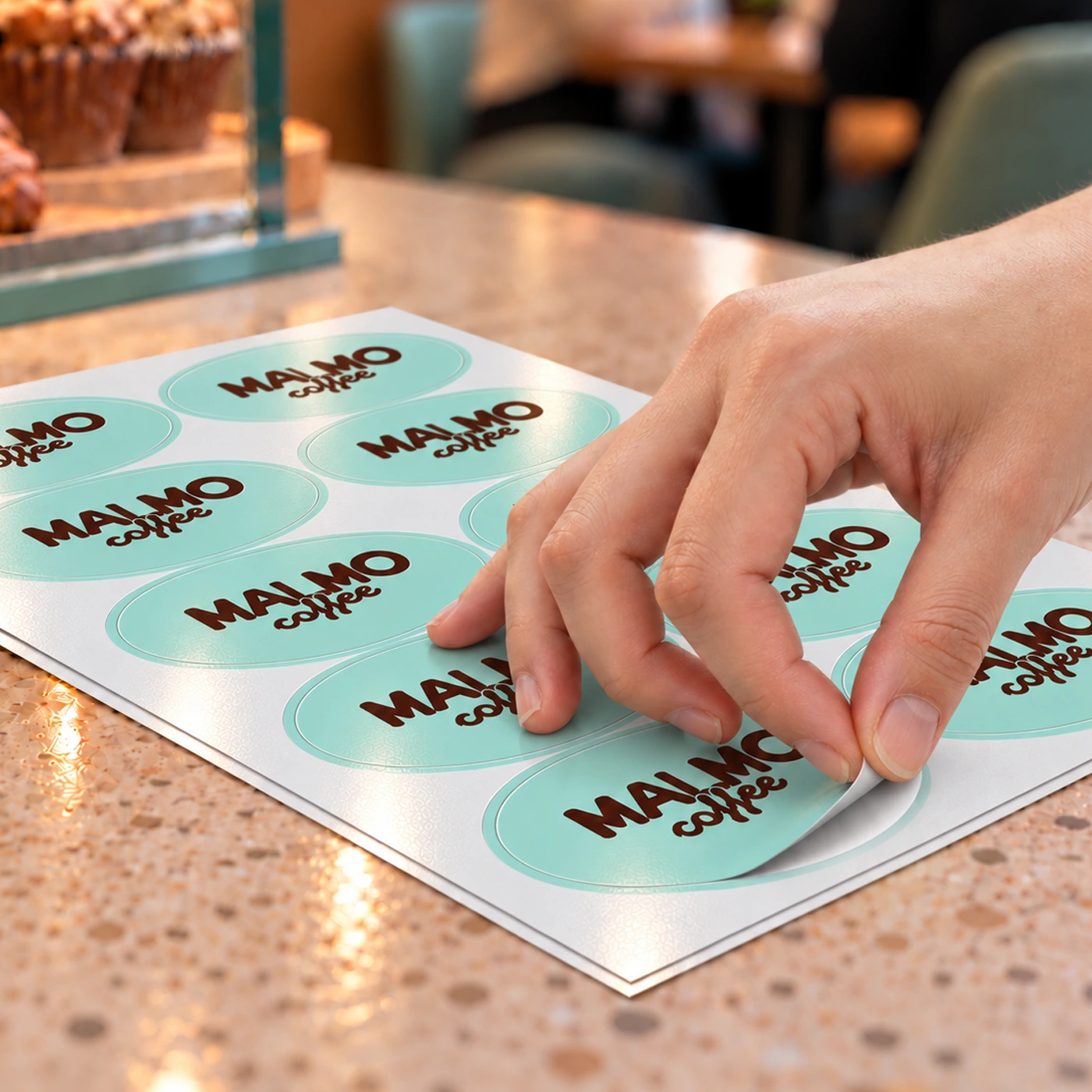 Applying a custom oval sticker from a branded sticker sheet in a cafe setting