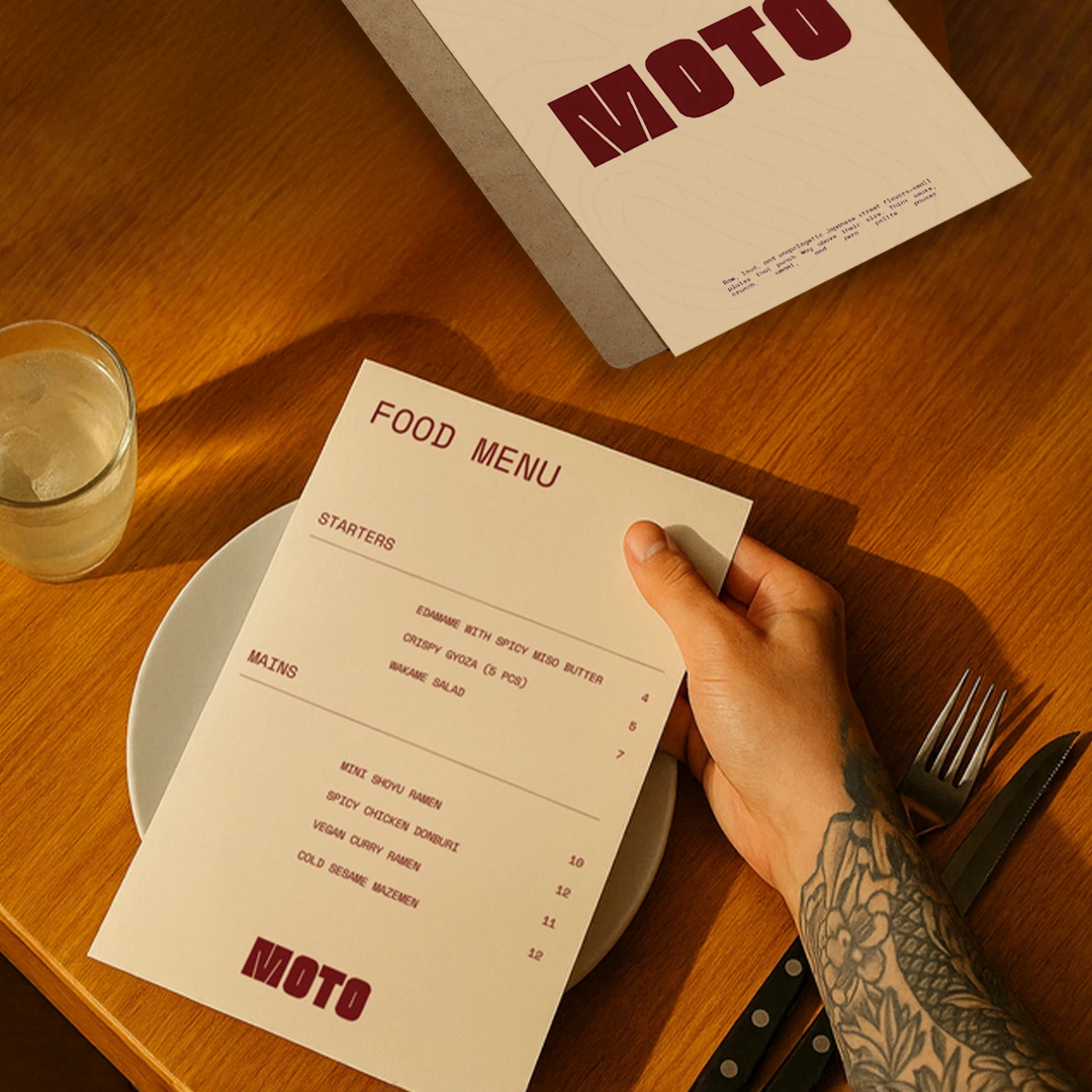A hand with a tattooed arm holds a modern "MOTO" food Menu over a wooden table set with a plate, cutlery, and a glass of water.