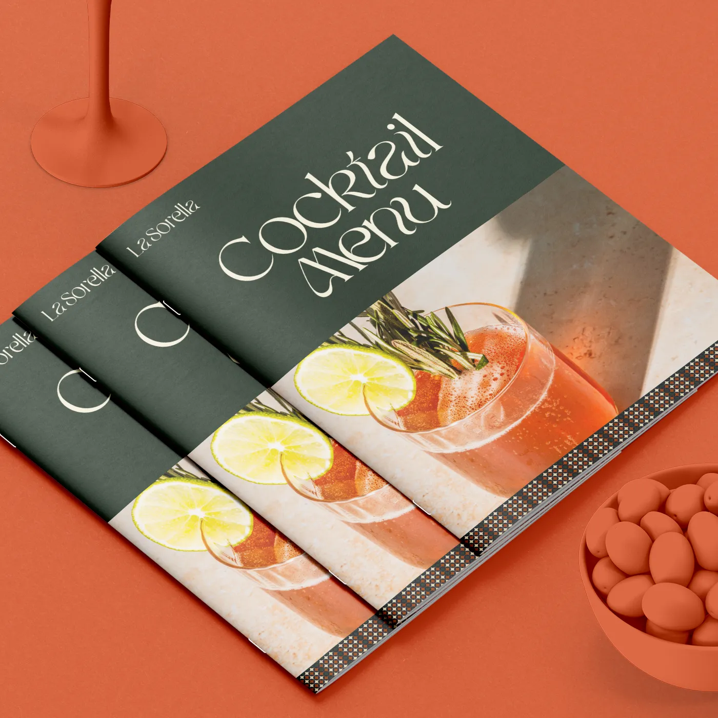 Three overlapping dark green cocktail Menu Booklets featuring a photo of a drink garnished with a lime wheel, resting on an orange surface.