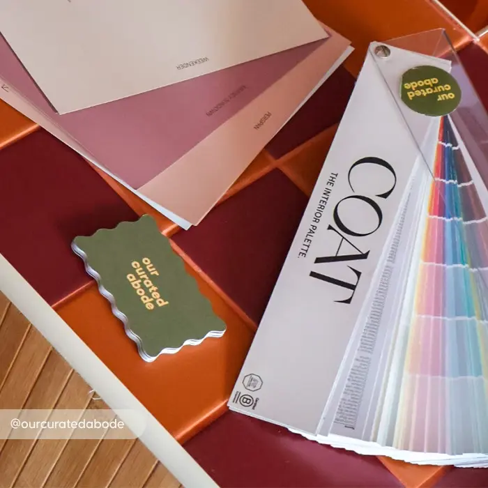 An olive green wavy shaped Business Card reading "our curated abode" beside a COAT paint fanning colour palette on a red and orange tiled surface.