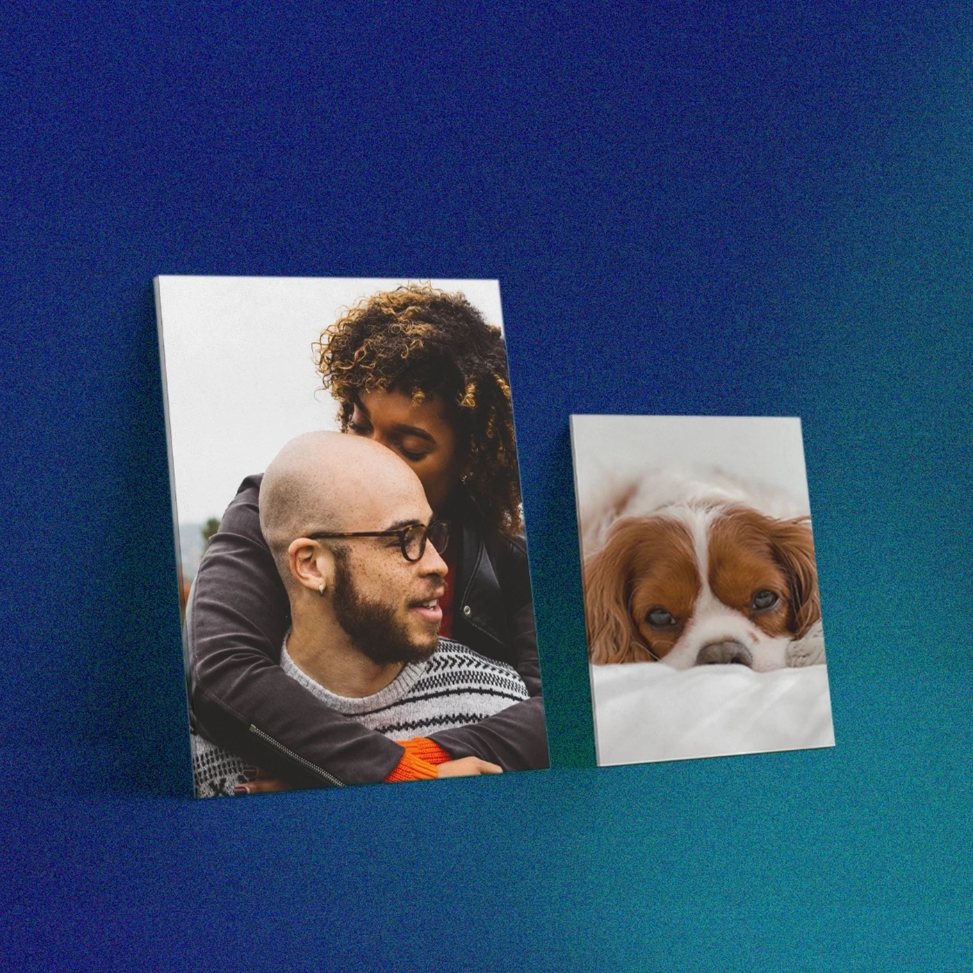 Two Christmas gifting Canvas Prints leaning against a dark blue wall: a larger print featuring a woman embracing a man from behind, and a smaller print showing a brown and white dog resting its head down.