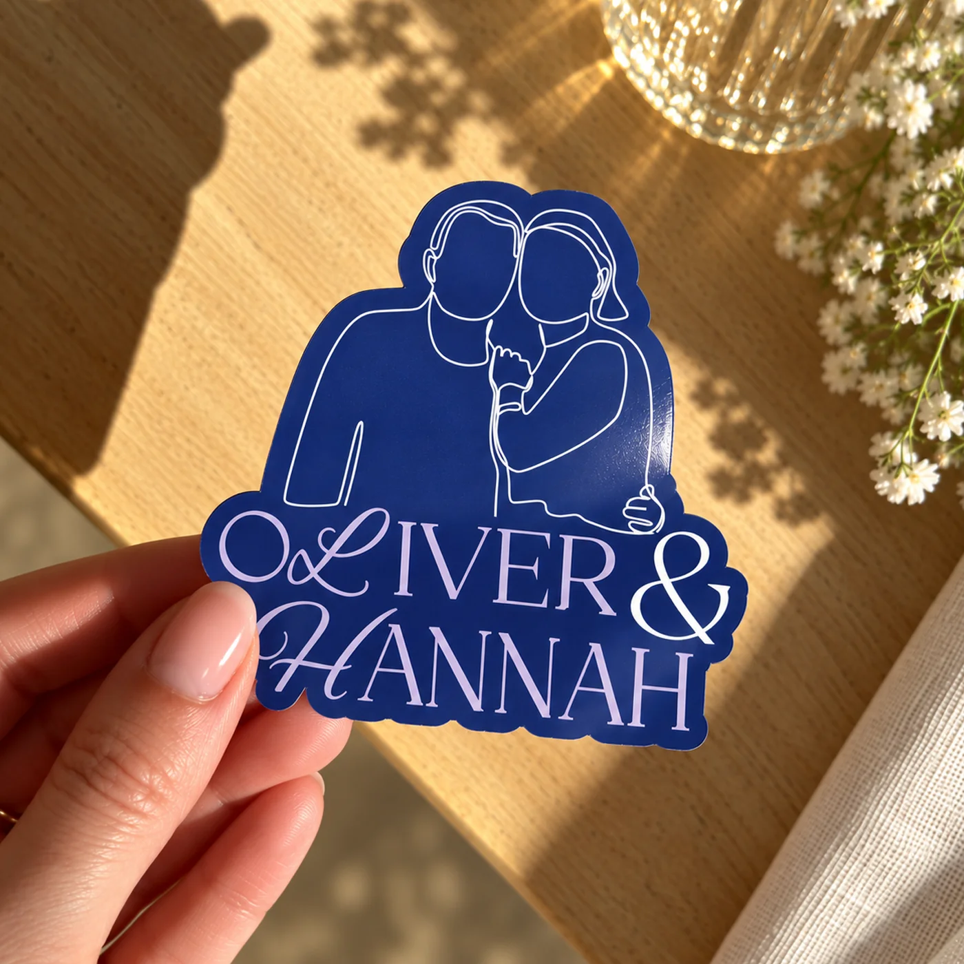 A hand holding a personalised dark blue Custom Shape Die Cut Sticker featuring a line-art illustration of a couple and the names "Oliver & Hannah".