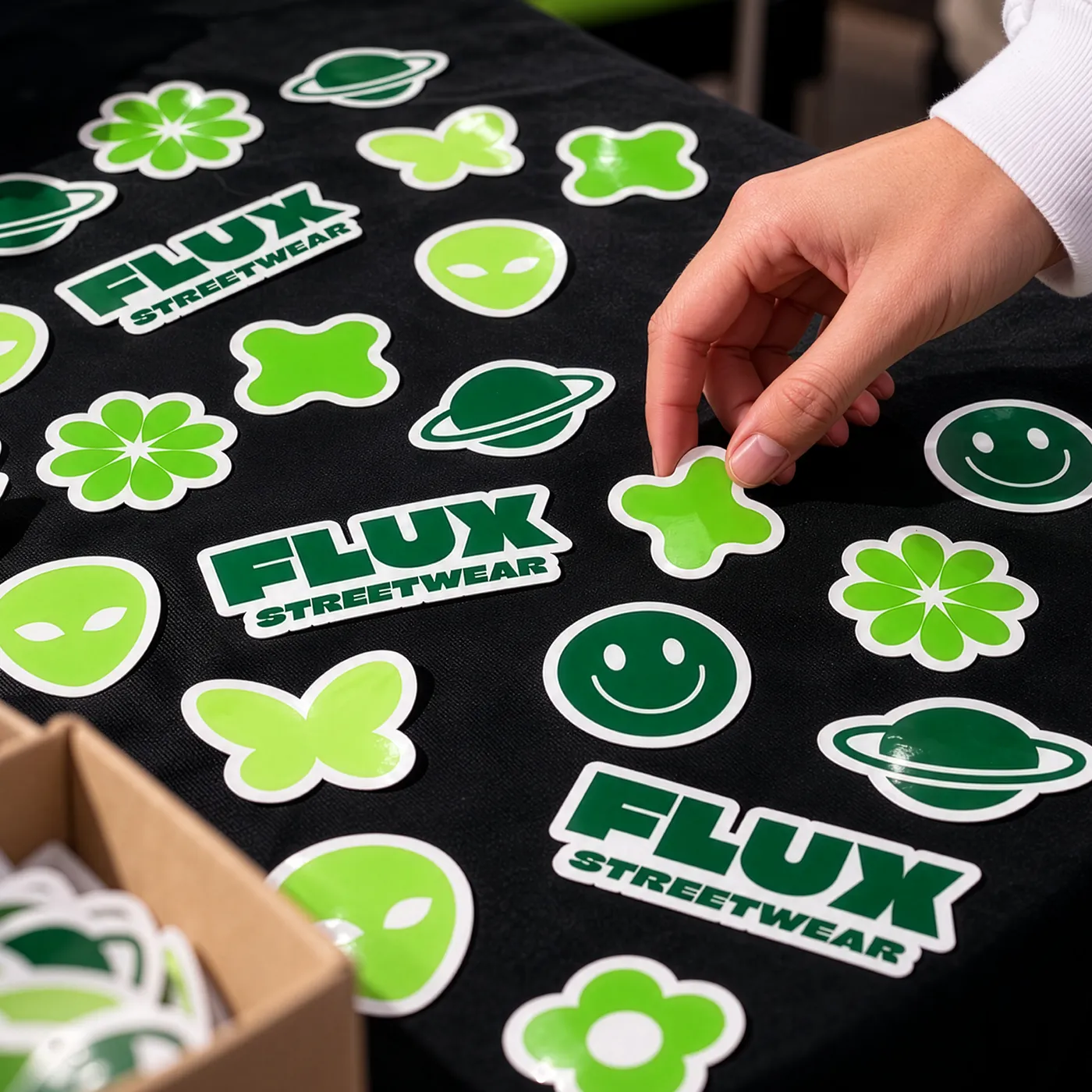 A hand picking up one of several green Custom Shape Die Cut streetwear Stickers, including aliens, planets, flowers, and FLUX logos.