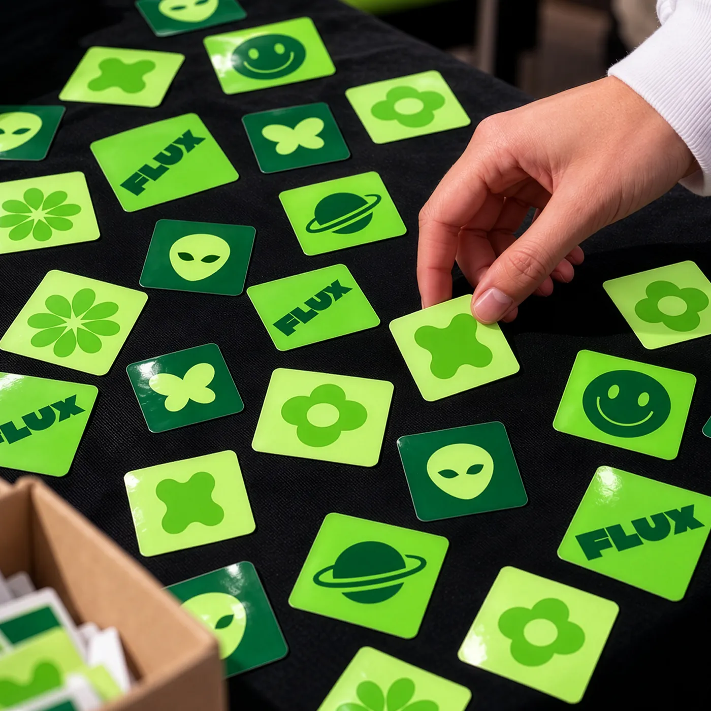 A hand picks up a bright green Square Die Cut Sticker among bold green smiley faces, clovers, flowers, alien and planet designs, and “FLUX” branding.