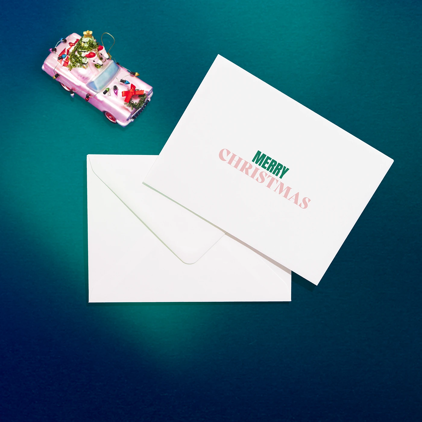 A white "MERRY CHRISTMAS" card and matching Envelope lie on a teal background near a toy pink car carrying a Christmas tree.