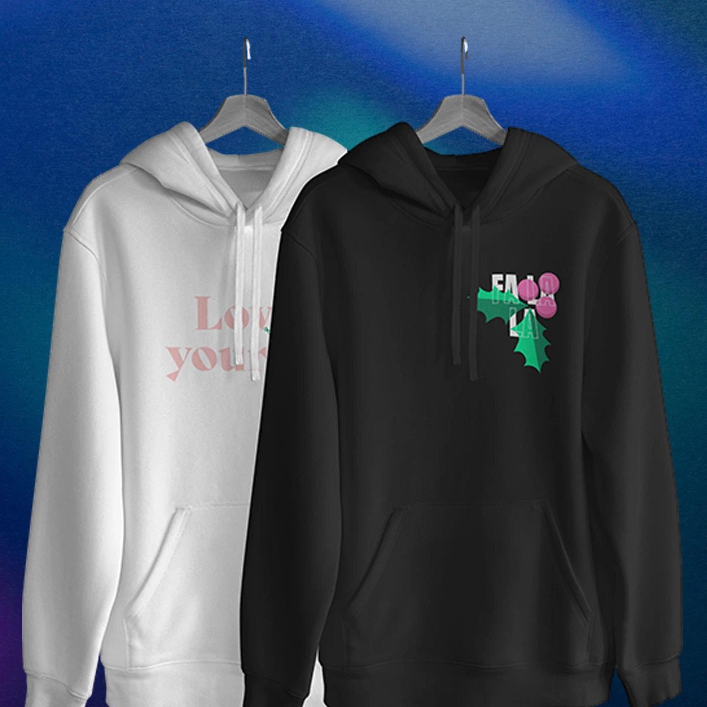 Two Christmas Hoodies hanging side by side on dark hangers against a blue background: a white hoodie on the left with pink text reading "Love yourself" and a black hoodie on the right featuring a green holly and pink berries graphic on the left chest.