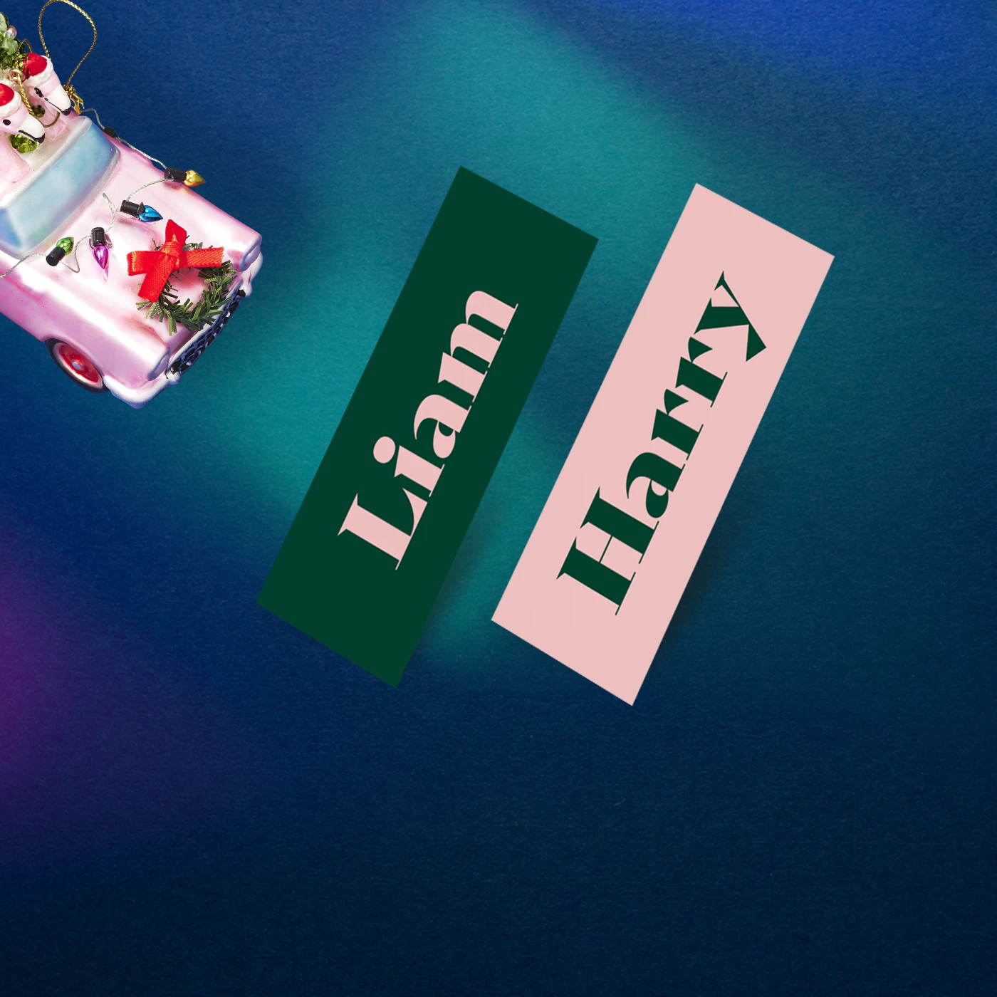 Two Christmas Mini Business Cards — one green with pink text reading "Liam" and one pink with green text reading "Harry"—lie on a blue background near a toy pink car decorated with a festive wreath.