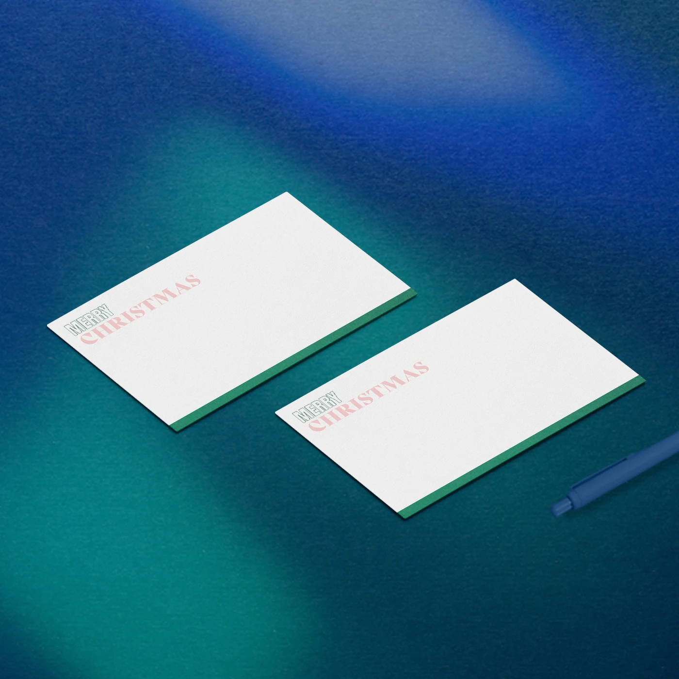 Two Christmas Notecards with green-edged bottoms and pink "MERRY CHRISTMAS" text resting on a dark blue background alongside a blue pen