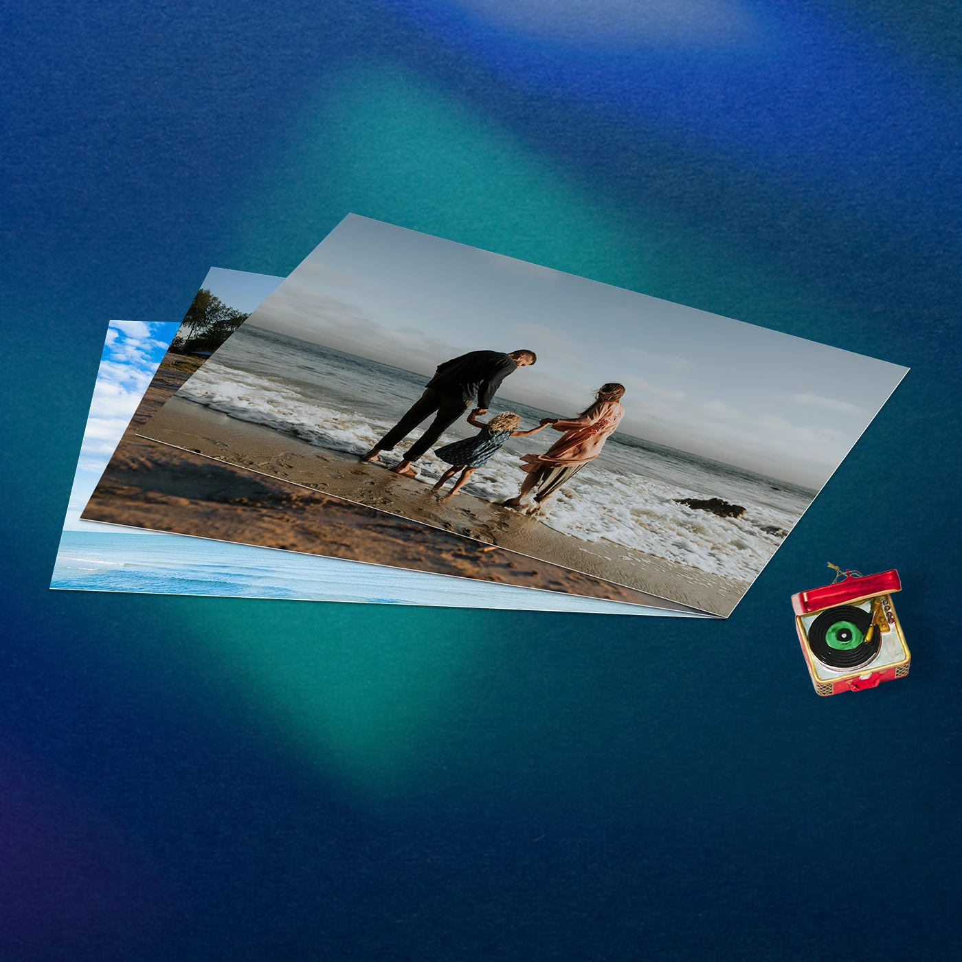 A stack of Christmas Photo Prints, topped by one of a family on the beach, rests on a blue surface next to a miniature record player charm.