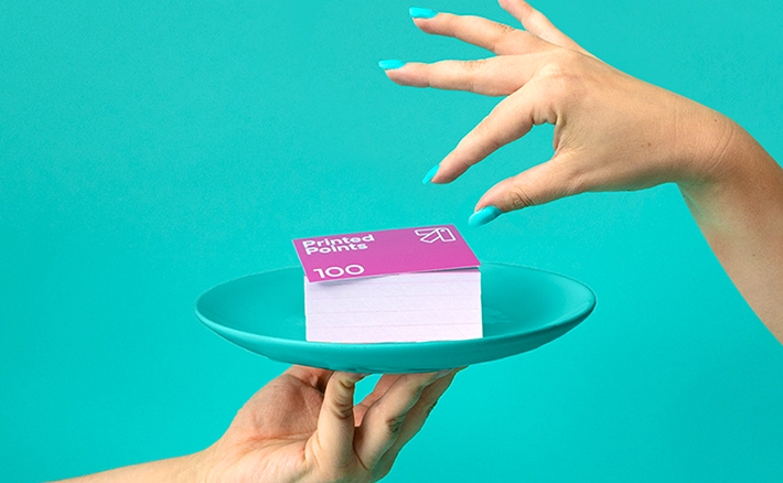 gainst a bright teal background, one hand holds out a matching teal plate carrying a stack of business cards topped with a magenta Printed Points 100 voucher, while another hand reaches down towards it.