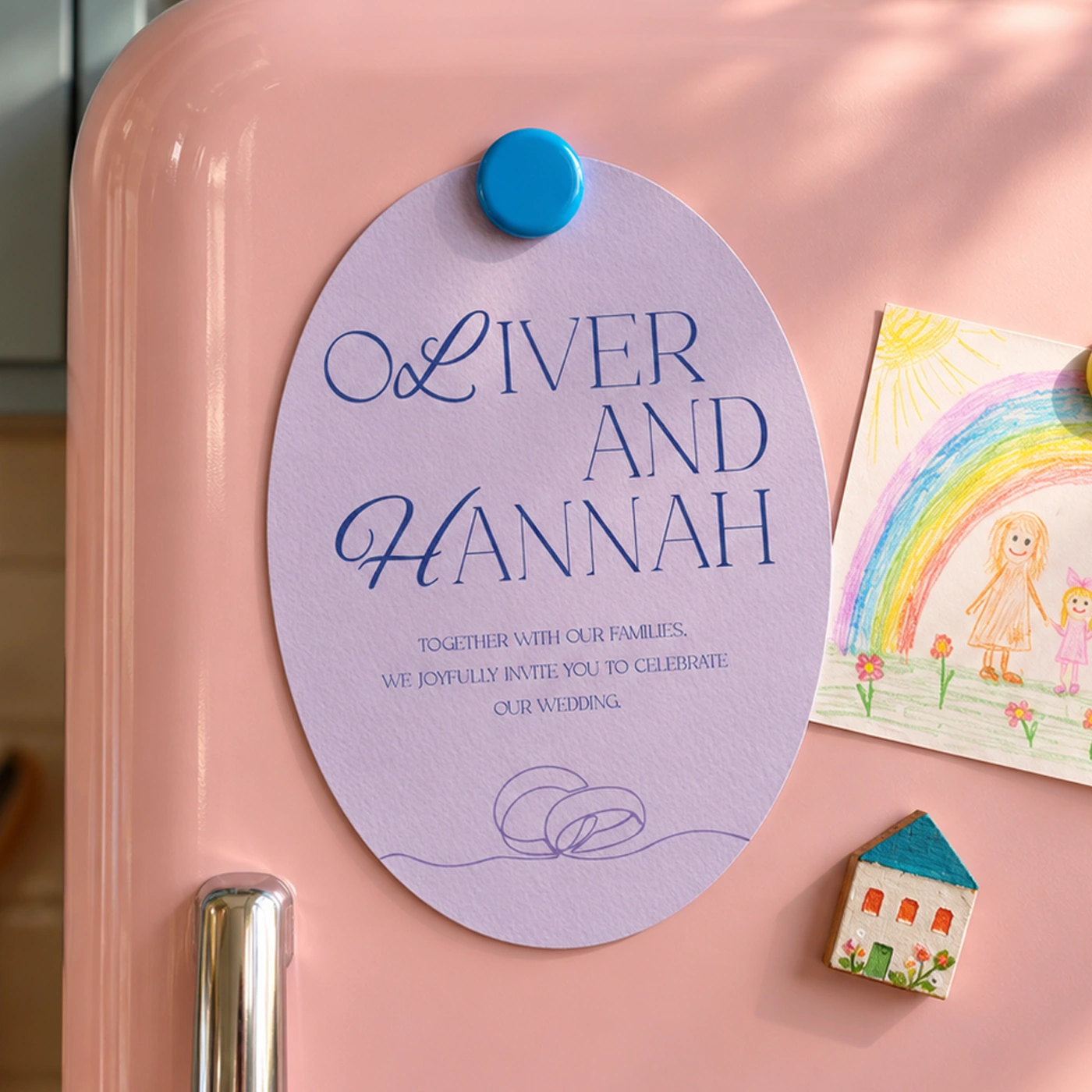 Lilac oval wedding invitation held on a pink fridge with a blue magnet, next to a child’s rainbow drawing