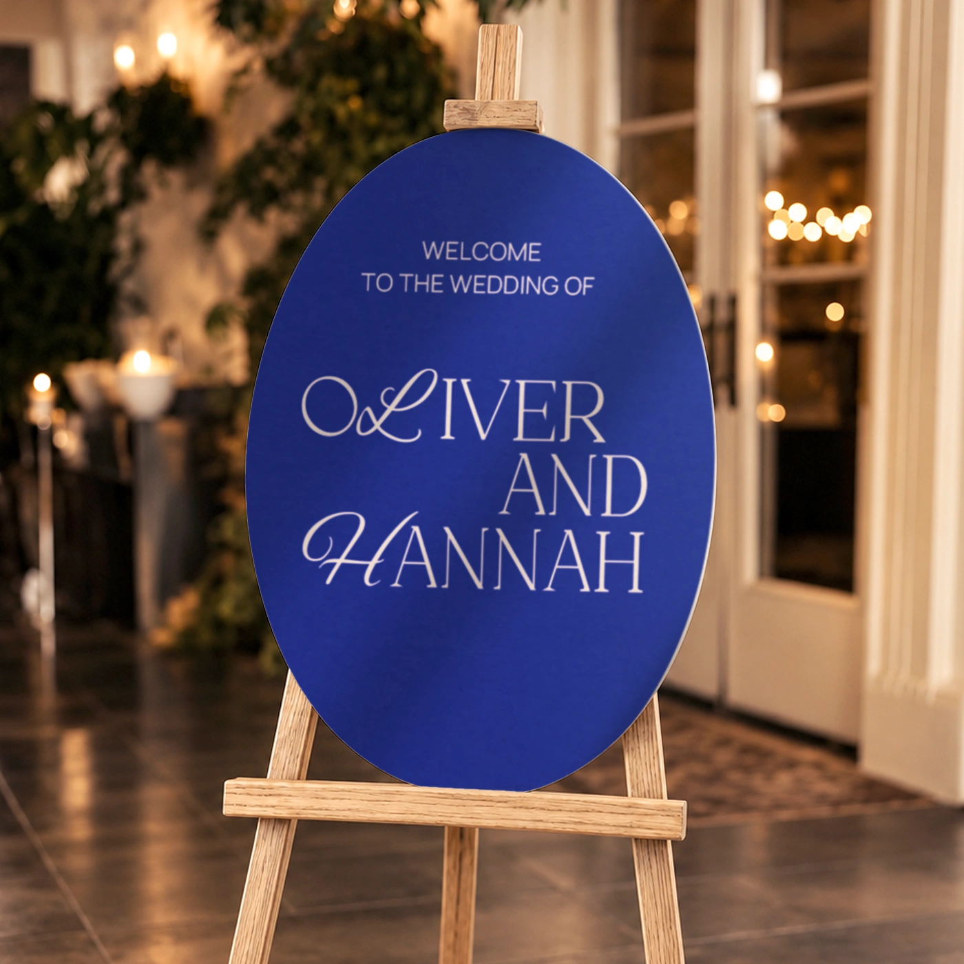 Blue oval wedding welcome sign with the couple’s names, displayed on a wooden easel at a venue entrance