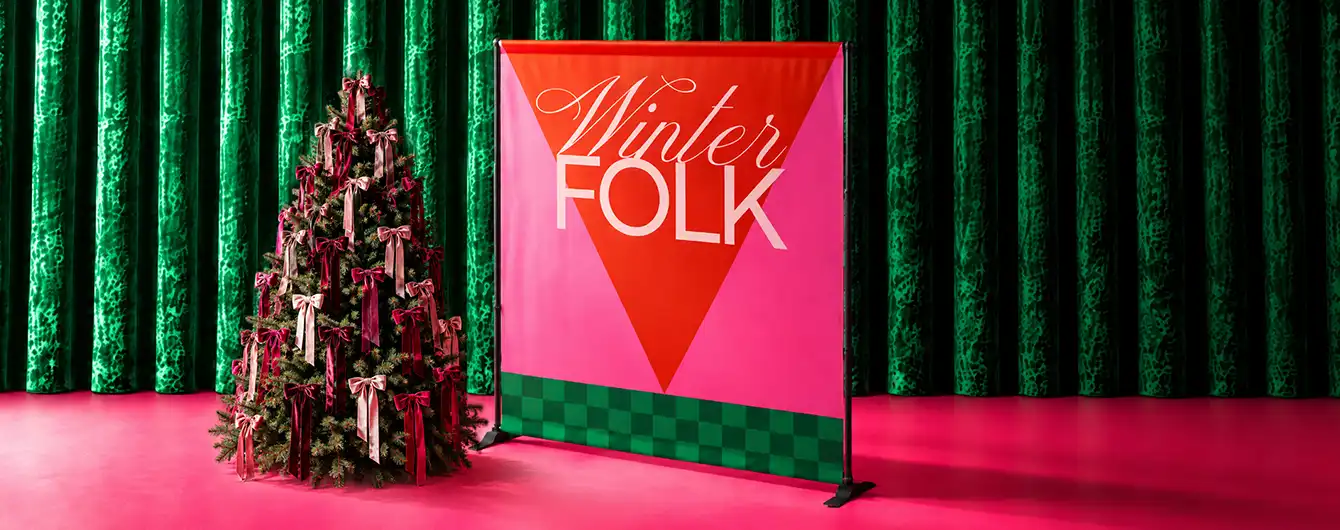 A large red and pink "Winter FOLK" Backdrop Banner set up next to a bow-decorated Christmas tree against a green velvet curtain background