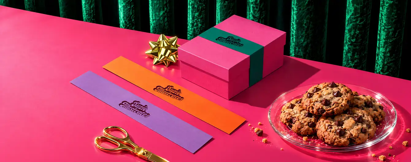 A bright pink gift box with a green Belly Band, displayed alongside colourful Belly Bands laid out, gold scissors, a gold bow, and a plate of chocolate chip cookies on a pink surface.